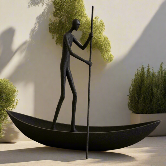 Black sculpture of a person in a boat with a pole against a light wall with greenery