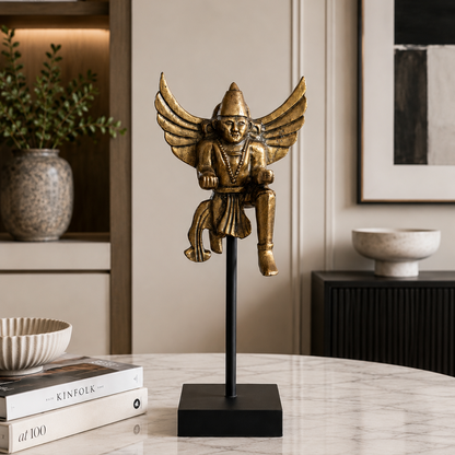 Gold angel figurine on a black stand in a home decor setting with books and a plant.